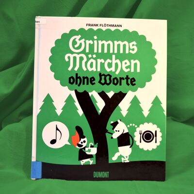 Foto des Buches "Grimms M&auml;rchen ohne Worte" vor gr&uuml;nem Hintergrund. Das Buchcover zeigt einen Baum, in dessen Krone der Buchtitel steht. Links neben dem Baumstamm steht ein Kind mit roter Kappe, in seiner Sprechblase erscheint eine Musiknote. Rechts neben dem Baumstamm steht ein Wolf, in dessen "Gedanken"-Sprechblase ein Teller mit Messer und Gabel abgebildet ist.