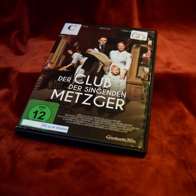 Foto der DVD "Der Club der singenden Metzger". Das Cover zeigt eine Familie, die sich wie f&uuml;r einen Fototermin aufstellt: Der Ehemann h&auml;lt ein Baby im Arm, vor der Ehefrau steht der kleine Sohn. Im Hintergrund sieht eine junge Frau die Familie an.