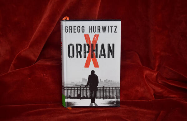 Foto des Romans "Orphan X": Das schwarzwei&szlig;e Cover zeigt die Silhouette eines Mannes und direkt &uuml;ber seinem Kopf als einzigen Farbakzent das gro&szlig;e rote "X" aus dem ansonsten in schwarz geschriebenen Buchtitel.