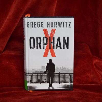 Foto des Romans "Orphan X": Das schwarzwei&szlig;e Cover zeigt die Silhouette eines Mannes und direkt &uuml;ber seinem Kopf als einzigen Farbakzent das gro&szlig;e rote "X" aus dem ansonsten in schwarz geschriebenen Buchtitel.