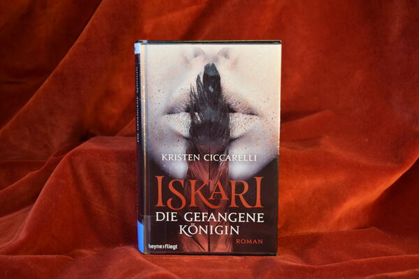 Foto des Buches "Die gefangene K&ouml;nigin". Der Hintergrund des Covers zeigt die untere H&auml;lfte eines Frauengesichts, das sich eine lange Feder senkrecht &uuml;ber die Lippen h&auml;lt.