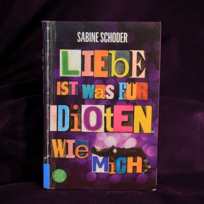 Foto des Buches "Liebe ist was f&uuml;r Idioten. Wie mich". Das Cover zeigt den Buchtitel wie einzeln nebeneinandergelegte Buchstaben aus verschiedenen Alphabeten und in unterschiedlichen Farben. Auf dem letzten Wort "mich" liegt ein M&auml;dchen in schwarzer Kleidung mit verschr&auml;nkten Armen.