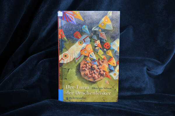 Foto des Buches "Der Turm der Drachenlenker". Das gezeichnete Cover zeigt aus der Vogelperspektive ein kleines Dorf, &uuml;ber dem viele bunte Stoffdrachen in unterschiedlichen Farben und Formen schweben.