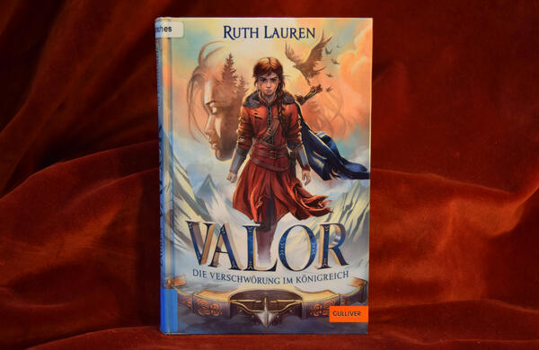 Foto des Buches "Valor - Die Verschw&ouml;rung im K&ouml;nigreich". Das gezeichnete Cover zeigt ein entschlossen voranschreitendes M&auml;dchen in Winterkleidung mit einem K&ouml;cher voller Pfeile &uuml;ber der Schulter, im Hintergrund ein M&auml;dchengesicht mit geschlossenen Augen und vorne am unteren Bildrand eine Armbrust mit eingelegtem Pfeil.