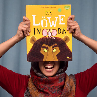 "Bookface"-Foto des Buches "Der L&ouml;we in dir". Das gezeichnete Cover zeigt eine kleine Maus, die auf dem Kopf eines L&ouml;wen steht. Vom Gesicht des L&ouml;wen ist nur die obere H&auml;lfte zu sehen. Das Buch wird von einer Person so gehalten, dass es die obere H&auml;lfte ihres Gesichts verdeckt, w&auml;hrend sie den Mund zu einem L&ouml;wenbr&uuml;llen &ouml;ffnet.