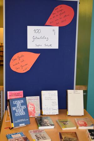 Foto eines mit B&uuml;chern best&uuml;ckten Tisches im Vordergrund, dahinter eine Pinnwand mit drei Beschriftungen: "100. Geburtstag Sophie Scholl" in der Mitte, umgeben von "Das Gesetz &auml;ndert sich. Das Gewissen nicht!" und "Steh zu den Dingen, an die du glaubst. Auch wenn du alleine dort stehst.".