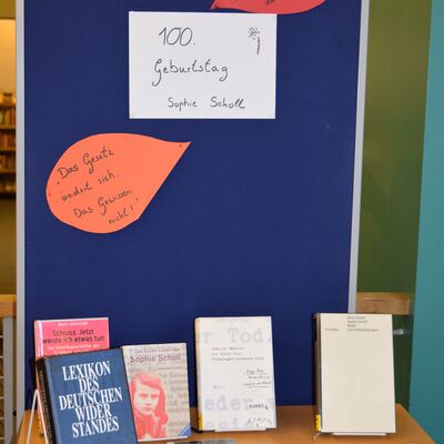 Foto eines mit B&uuml;chern best&uuml;ckten Tisches im Vordergrund, dahinter eine Pinnwand mit drei Beschriftungen: "100. Geburtstag Sophie Scholl" in der Mitte, umgeben von "Das Gesetz &auml;ndert sich. Das Gewissen nicht!" und "Steh zu den Dingen, an die du glaubst. Auch wenn du alleine dort stehst.".