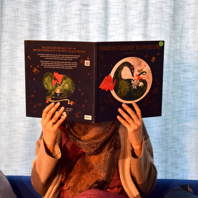 Vor einem hellblauen Vorhang sitzt eine Person und liest aus dem gro&szlig;en Bilderbuch "Franklins fliegende Buchhandlung" vor. Ihr Gesicht wird durch das aufgeklappte Buch verdeckt.