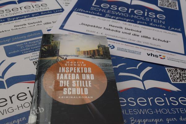 Foto des Buches "Inspektor Takeda und die stille Schuld". Im Hintergrund sind mehrere Plakate der "Lesereise Schleswig-Holstein" mit der Ank&uuml;ndigung f&uuml;r die Lesung zu sehen.