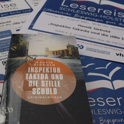 Foto des Buches "Inspektor Takeda und die stille Schuld". Im Hintergrund sind mehrere Plakate der "Lesereise Schleswig-Holstein" mit der Ank&uuml;ndigung f&uuml;r die Lesung zu sehen.