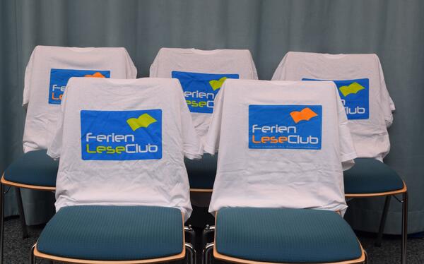 Foto von f&uuml;nf St&uuml;hlen, &uuml;ber deren R&uuml;ckenlehnen jeweils wei&szlig;e T-Shirts mit aufgedrucktem FerienLeseClub-Logo gezogen wurden.