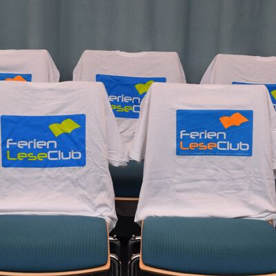 Foto von f&uuml;nf St&uuml;hlen, &uuml;ber deren R&uuml;ckenlehnen jeweils wei&szlig;e T-Shirts mit aufgedrucktem FerienLeseClub-Logo gezogen wurden.