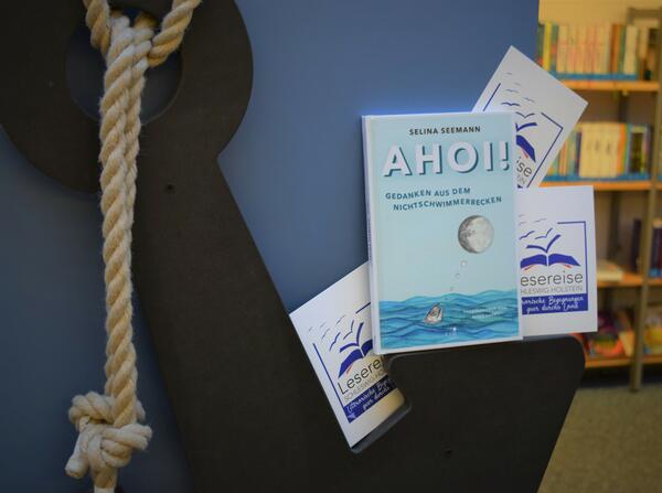 Foto des Buches "Ahoi!" von Selina Seemann zusammen mit Postkarten, auf denen das Logo der "Lesereise Schleswig-Holstein" abgebildet ist, platziert auf einem h&ouml;lzernen Anker.