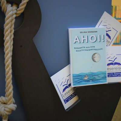 Foto des Buches "Ahoi!" von Selina Seemann zusammen mit Postkarten, auf denen das Logo der "Lesereise Schleswig-Holstein" abgebildet ist, platziert auf einem h&ouml;lzernen Anker.