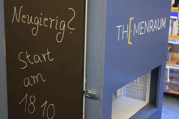 Nahaufnahme des M&ouml;belst&uuml;cks "Themenraum": Zu sehen sind links eine schwarze Tafel mit der wei&szlig;en Aufschrift "Neugierig? Start am 18.10." und rechts eine bl&auml;uliche, noch geschlossene T&uuml;r mit der Aufschrift "Themenraum".