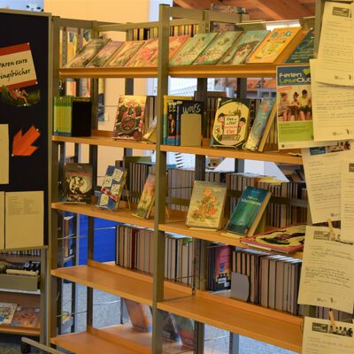 Foto des Ausstellungsregals, in dem einige der beliebtesten FerienLeseClub-B&uuml;cher des Jahres 2021 gezeigt werden. Links steht eine Stellwand, auf der die mehrseitige Liste der beliebtesten Titel angebracht ist. Rechts am Regal ist eine Auswahl von bearbeiteten FLC-Kreativb&ouml;gen und FLC-Frageb&ouml;gen zu sehen.