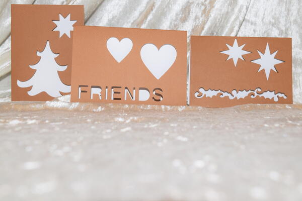 Foto von drei hellbraunen Klappkarten mit jeweils unterschiedlichen ausgeschnittenen Motiven zum Thema Freundschaft und Weihnachten in wei&szlig;glitzernder Umgebung.