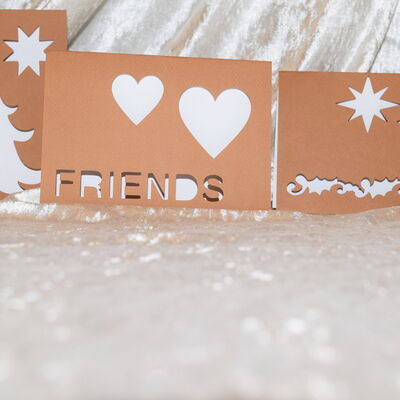 Foto von drei hellbraunen Klappkarten mit jeweils unterschiedlichen ausgeschnittenen Motiven zum Thema Freundschaft und Weihnachten in wei&szlig;glitzernder Umgebung.