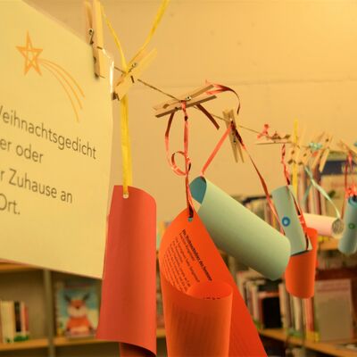 Foto einer W&auml;scheleine, an der bunte zusammengerollte Papiere mit B&auml;ndern und Klammern aufgeh&auml;ngt sind. Links im Vordergrund ein Infozettel mit der Beschriftung "Pfl&uuml;ck dir ein Weihnachtsgedicht und gib es weiter oder h&auml;nge es bei dir Zuhause an einen sch&ouml;nen Ort."
