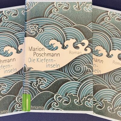 Foto von drei Exemplaren des Buches "Die Kieferninseln" von Marion Poschmann, die f&auml;cherf&ouml;rmig &uuml;bereinander gelegt sind. Das Cover zeigt stilisierte Meereswogen in blau-wei&szlig;.