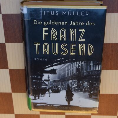 Foto des Buches "Die goldenen Jahre des Franz Tausend" auf einem Schachbrett. Das Cover zeigt eine alte Schwarz-Wei&szlig;-Aufnahme aus dem Stra&szlig;enverkehr einer gro&szlig;en Stadt; dar&uuml;ber steht der Buchtitel in goldener Schrift.