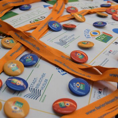 Foto von Bronze-, Silber- und Gold-Urkunden f&uuml;r den FerienLeseClub zusammen mit FerienLeseClub-Schl&uuml;sselb&auml;ndern und verschiedenfarbigen Buttons mit dem neuen FerienLeseClub-Logo