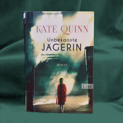 Foto des Buches "Unbekannte J&auml;gerin" von Kate Quinn. Das Cover zeigt eine Frau in rotem Mantel, deren Gesicht im Schatten kaum zu erkennen ist.