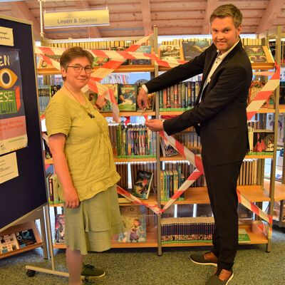 Das Foto zeigt das abgesperrte FerienLeseClub-Regal der Bibliothek sowie Petra Herzig und Sven Jensen beim Durchschneiden des Absperrbandes.